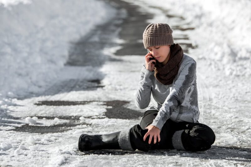 woman sitting on icy sidewalk holding leg while on phone