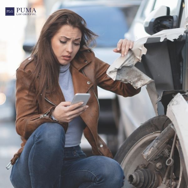 A woman in a brown jacket looks at her phone while inspecting damage to the front fender of a white car, possibly considering her next steps after an accident. The Puma Law Group logo appears in the upper left corner, highlighting a trusted car accident lawyer in Commack.